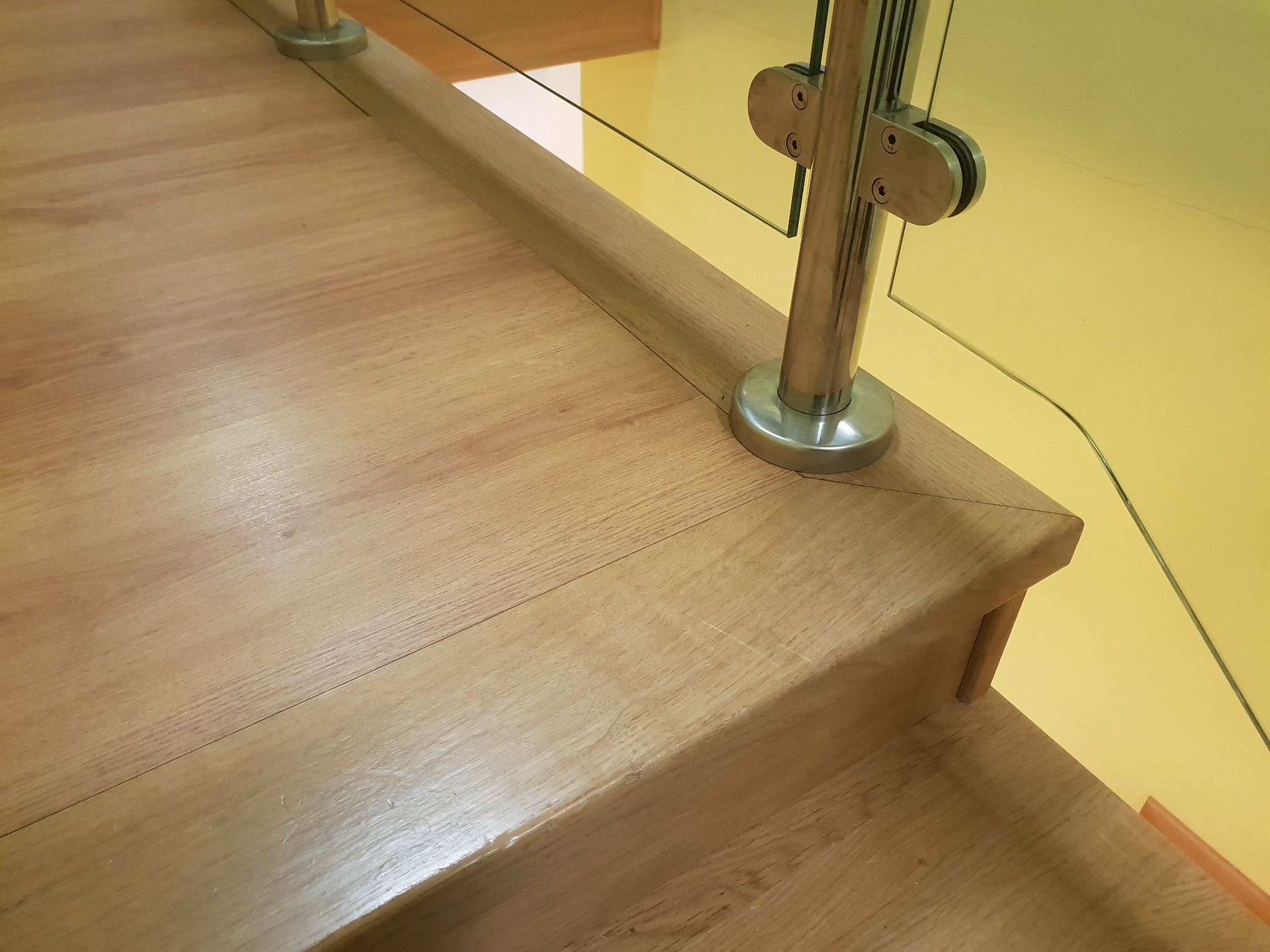 Fragment drewnianych schodów z balustradą ze szkła i metalu, widoczny detal wykończenia stopnia i mocowanie szkła.