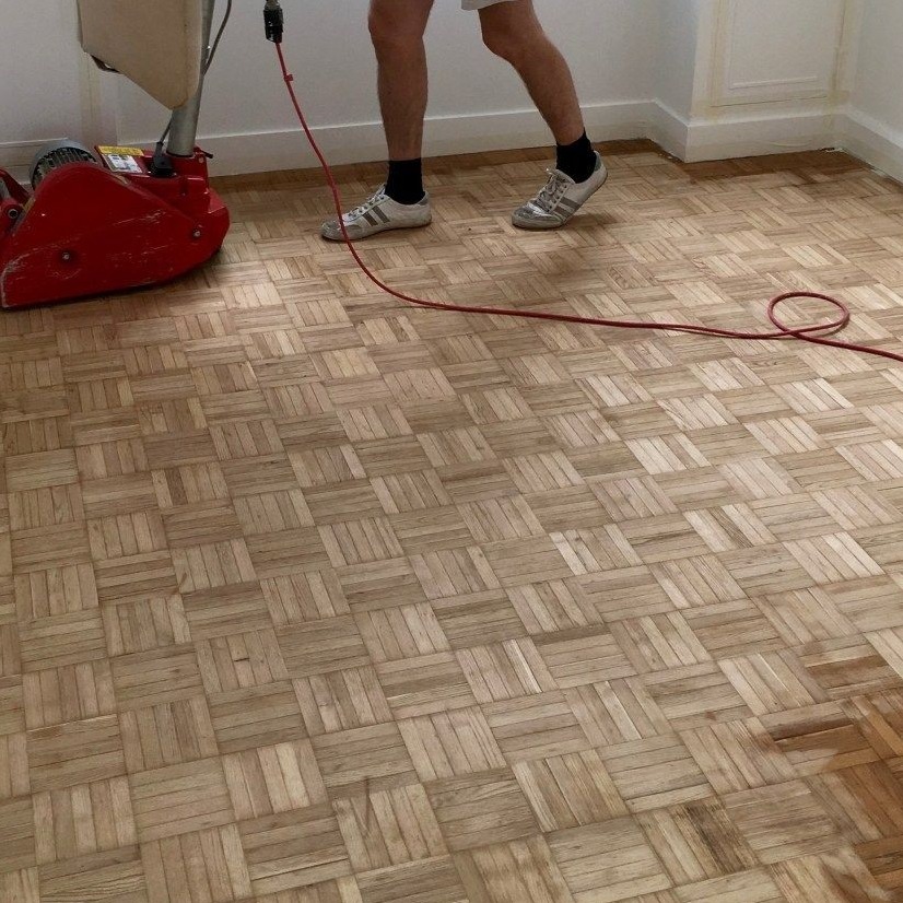 Szlifowanie parkietu ułożonego w kwadraty maszyną z czerwonym przewodem, widoczne nogi operatora w sportowych butach.