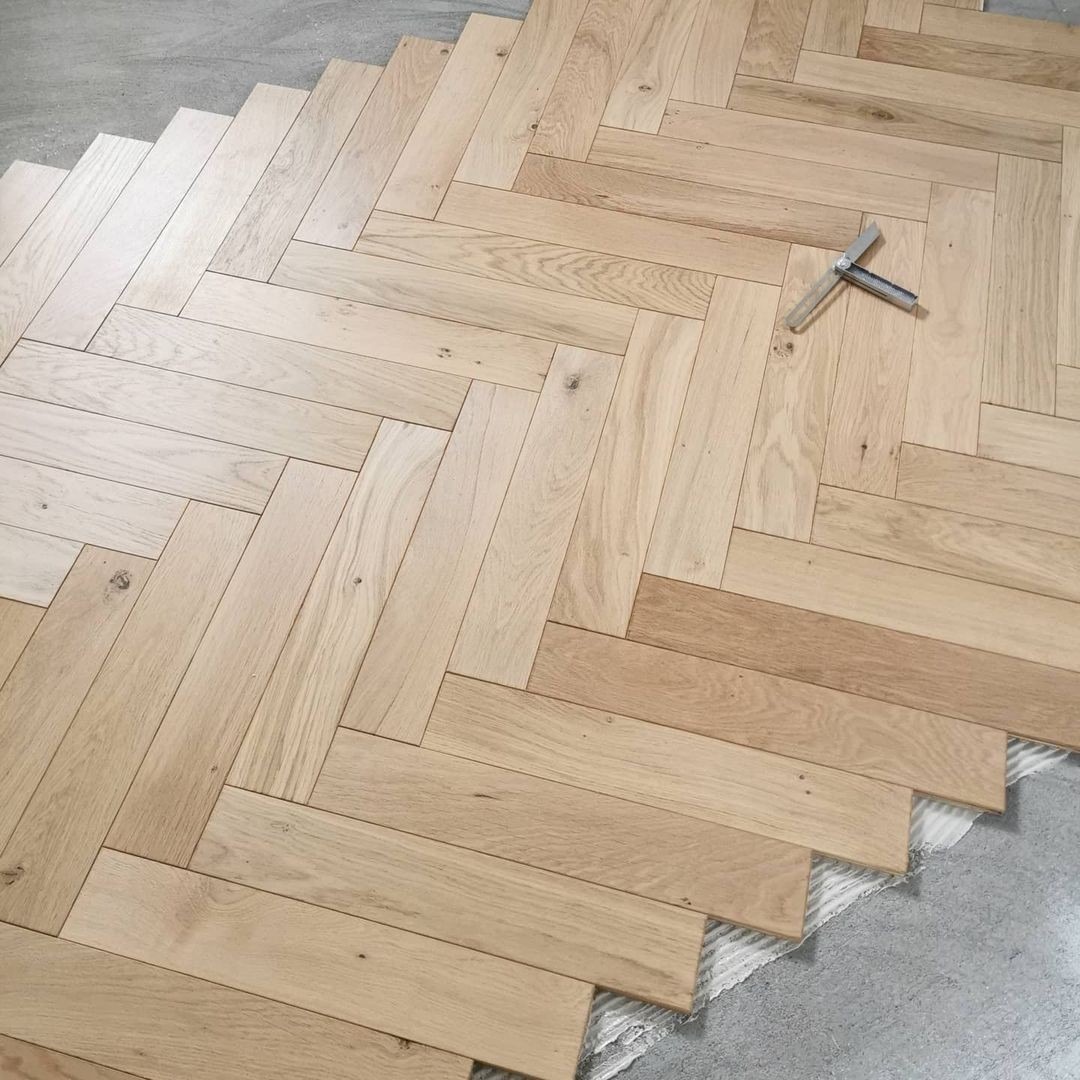 Jasne, dębowe deski ułożone w jodełkę na podłodze, widoczny fragment kleju montażowego i narzędzie do dociskania paneli.