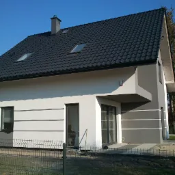 Nowoczesny dom jednorodzinny z jasną elewacją, ciemną dachówką i szarymi akcentami dekoracyjnymi na ścianach, widoczny zza metalowego ogrodzenia w słoneczny dzień.