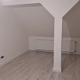 Poddasze po remoncie z panelami podłogowymi w odcieniach szarości, świeżo pomalowane na biało ściany i sufit z widoczną kolumną konstrukcyjną oraz grzejnikami pod skosem