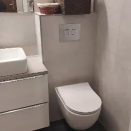 Nowoczesna toaleta podwieszana z szarą płytką imitującą kamień, białą umywalką nablatową i ciemną podłogą z płytek, uzupełniona o wiklinowy koszyk z ręcznikami.