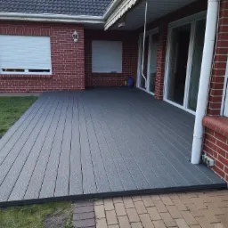 Szara deska kompozytowa tworząca taras przy ceglanym domu z roletami zewnętrznymi i częściowo widocznym trawnikiem.