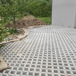 Ułożona nawierzchnia z ażurowych betonowych płyt na podjeździe, obrzeże z kamienia, widoczny fragment budynku z brązową bramą garażową i sterta ziemi w tle.