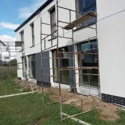 Dwukondygnacyjny dom z białą elewacją w trakcie prac wykończeniowych, widoczne rusztowania i fragmenty szarej elewacji, niezagospodarowany teren wokół budynku.
