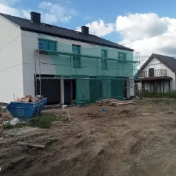 Nowy dom w trakcie prac elewacyjnych: biała fasada, rusztowania z zieloną siatką, kontener na gruz, teren budowy przed budynkiem.