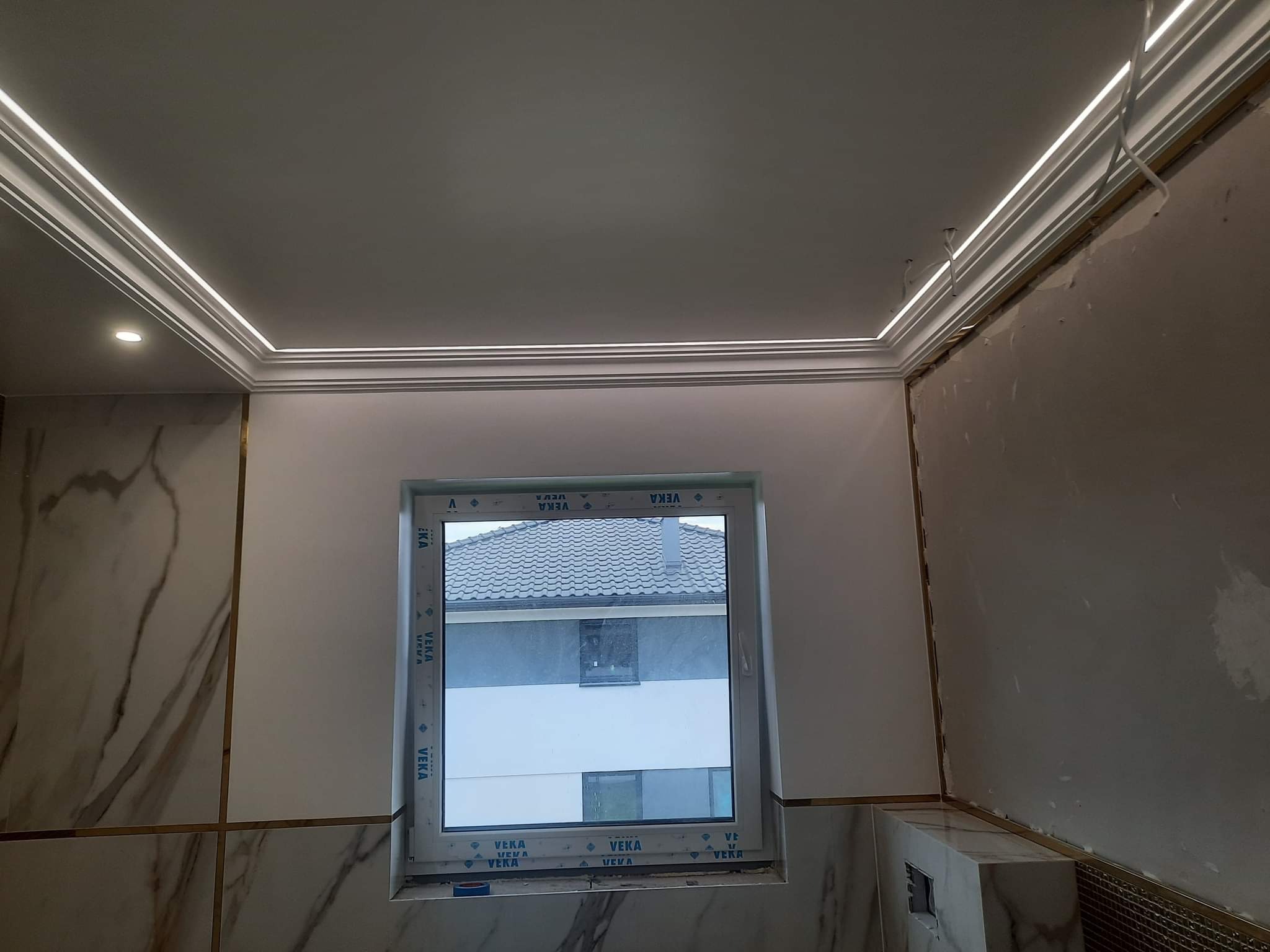 Fragment wnętrza w trakcie prac wykończeniowych: marmurowe płytki z złotymi listwami, okno PCV z widokiem na dom, sufit z oświetleniem LED w listwach, ściany częściowo przygotowane do malowania.