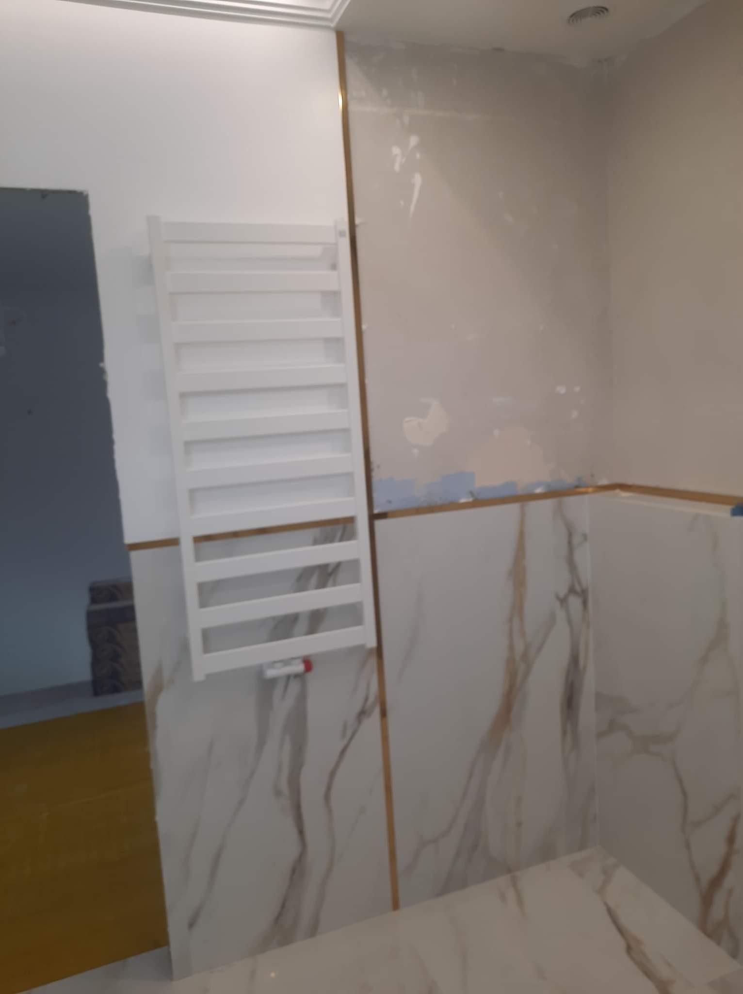 Wnętrze łazienki w trakcie prac: biały grzejnik drabinkowy na ścianie z marmurową okładziną, widoczne ślady po łataniu ścian, złote listwy dekoracyjne, żółta folia ochronna na podłodze w przejściu...