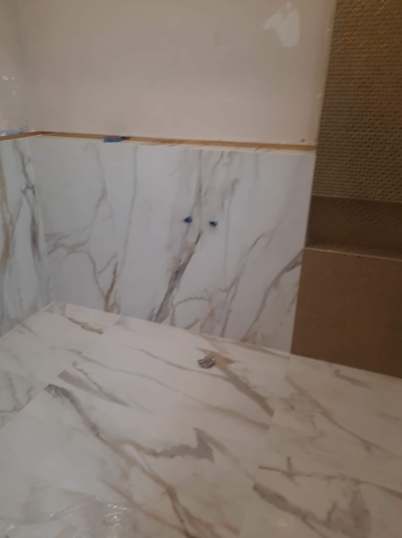 Układanie białych płytek imitujących marmur na podłodze i ścianach łazienki, widoczne niebieskie zaślepki na rurach.