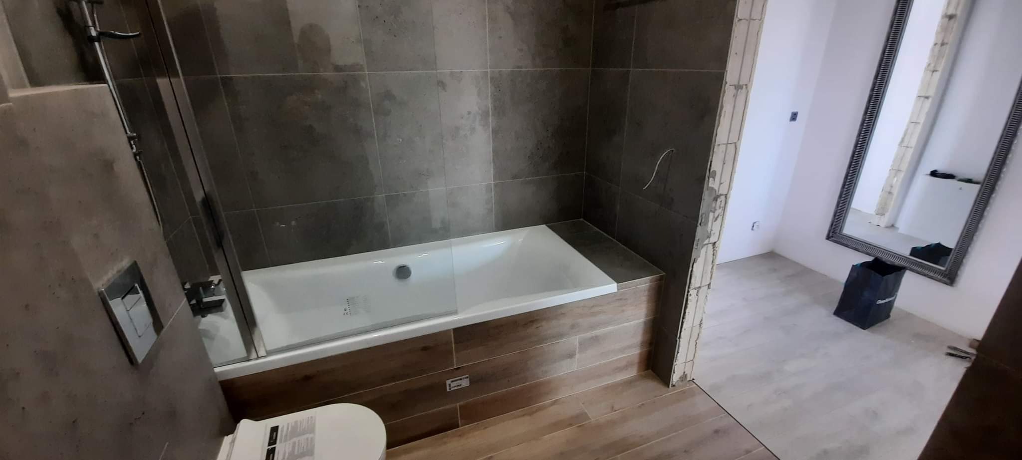 Nowoczesna łazienka w trakcie wykańczania: szare płytki na ścianach, obudowana wanna z drewnopodobnymi panelami, częściowo zamontowana toaleta, duże lustro w ozdobnej ramie i widoczne ślady prac...