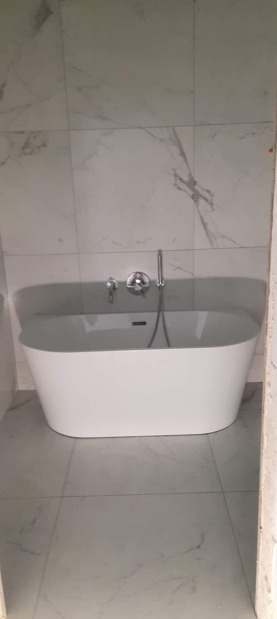 Nowoczesna, wolnostojąca wanna owalna z chromowaną armaturą na tle marmurowych płytek ściennych i podłogowych, widok z niskiej perspektywy w trakcie prac wykończeniowych.