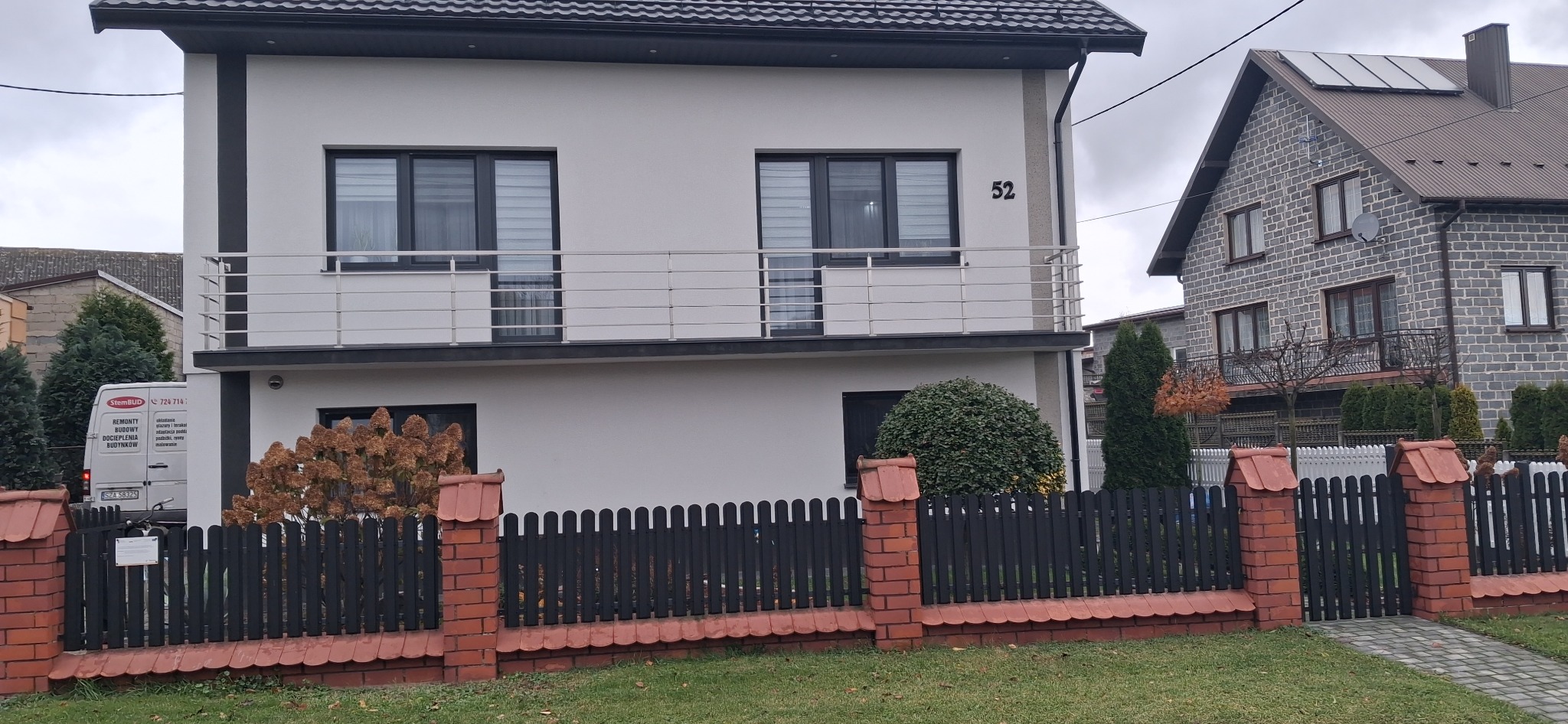 Dwupiętrowy dom z białymi ścianami i balkonem z metalową balustradą. Widoczny numer domu '52' i fragment ogrodzenia z cegły i czarnych sztachet.
