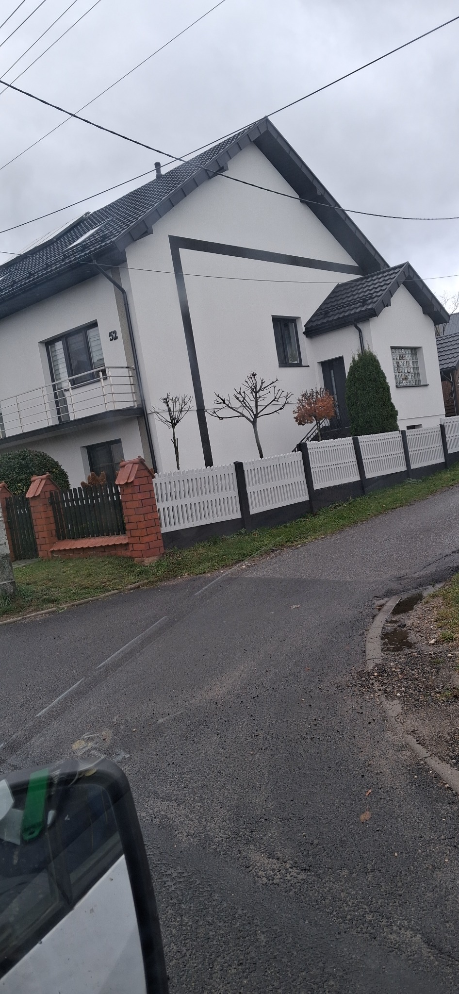 Biały dom z czarnymi akcentami i numerem 52, otoczony białym płotem z czarną podstawą. Widok z ulicy w pochmurny dzień.