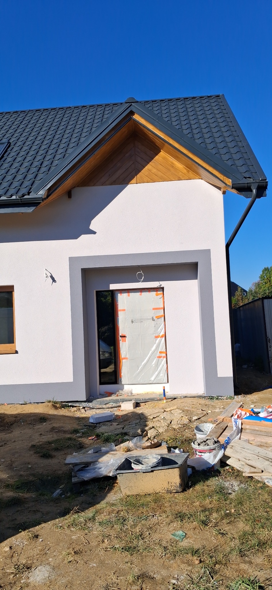 Jasny dom z ciemnym dachem i szarymi obramowaniami okien. Drzwi zabezpieczone folią. Widoczne materiały budowlane na ziemi. Błękitne niebo w tle.