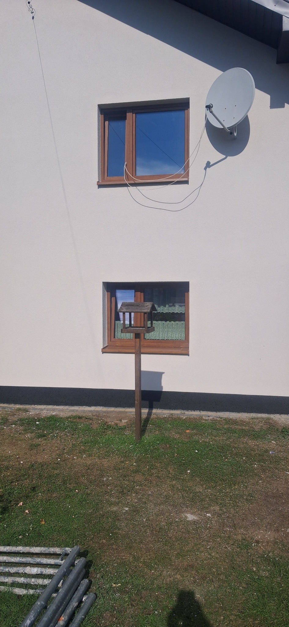 Jasna elewacja domu z brązowymi oknami i anteną satelitarną. Na trawniku karmnik dla ptaków. Widoczne rury na ziemi.