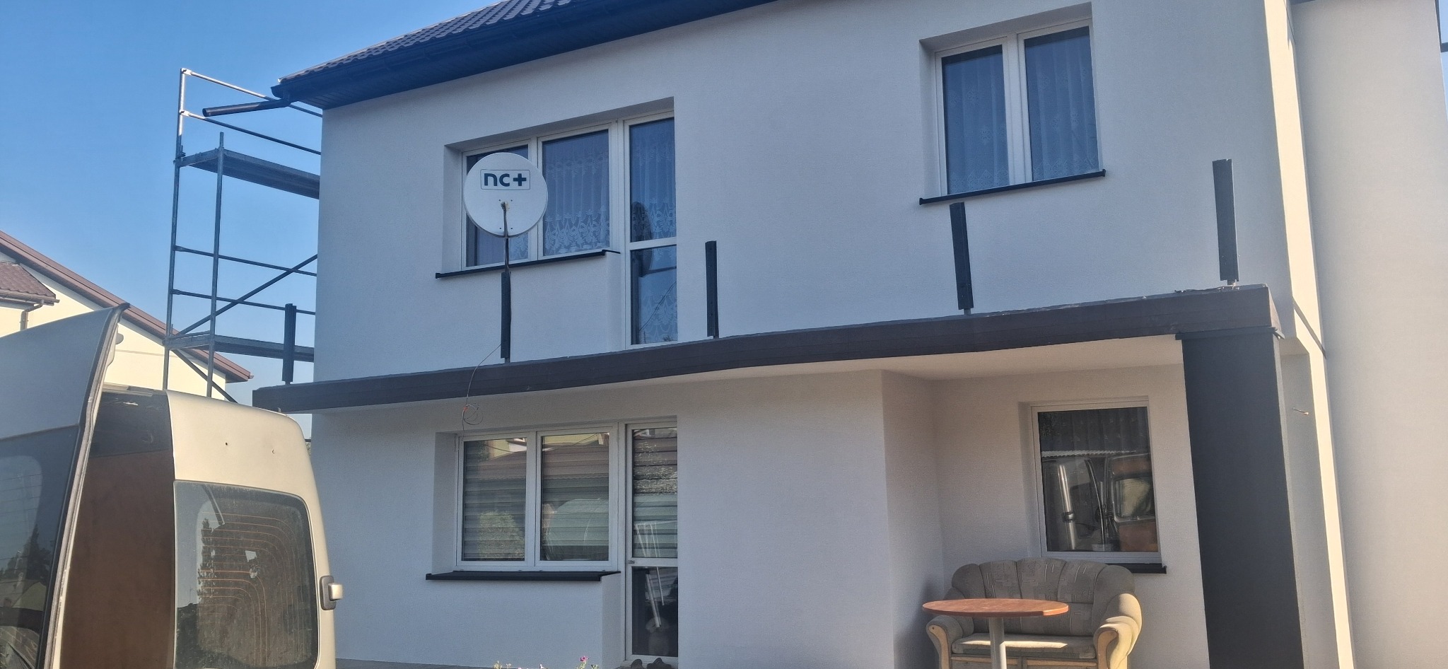 Biała elewacja domu po malowaniu, widoczne okna, antena satelitarna, rusztowanie z boku i częściowo zasłaniający kadr samochód dostawczy.