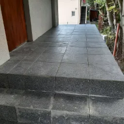 Zewnętrzne schody i taras wyłożone ciemnymi, kwadratowymi płytkami z delikatnym wzorem, prowadzące do drzwi wejściowych budynku.