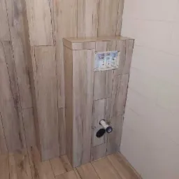 Obudowa stelaża WC z drewnopodobnych płytek ceramicznych, z widocznym otworem rewizyjnym i przyłączami kanalizacyjnymi, na tle ściany wyłożonej białymi płytkami i podłogi z drewnopodobnych paneli.