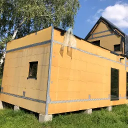 Surowy, żółty budynek w trakcie budowy, osadzony na betonowych blokach, z widocznymi oknami i czarną folią na dachu, otoczony zielenią i drzewami.