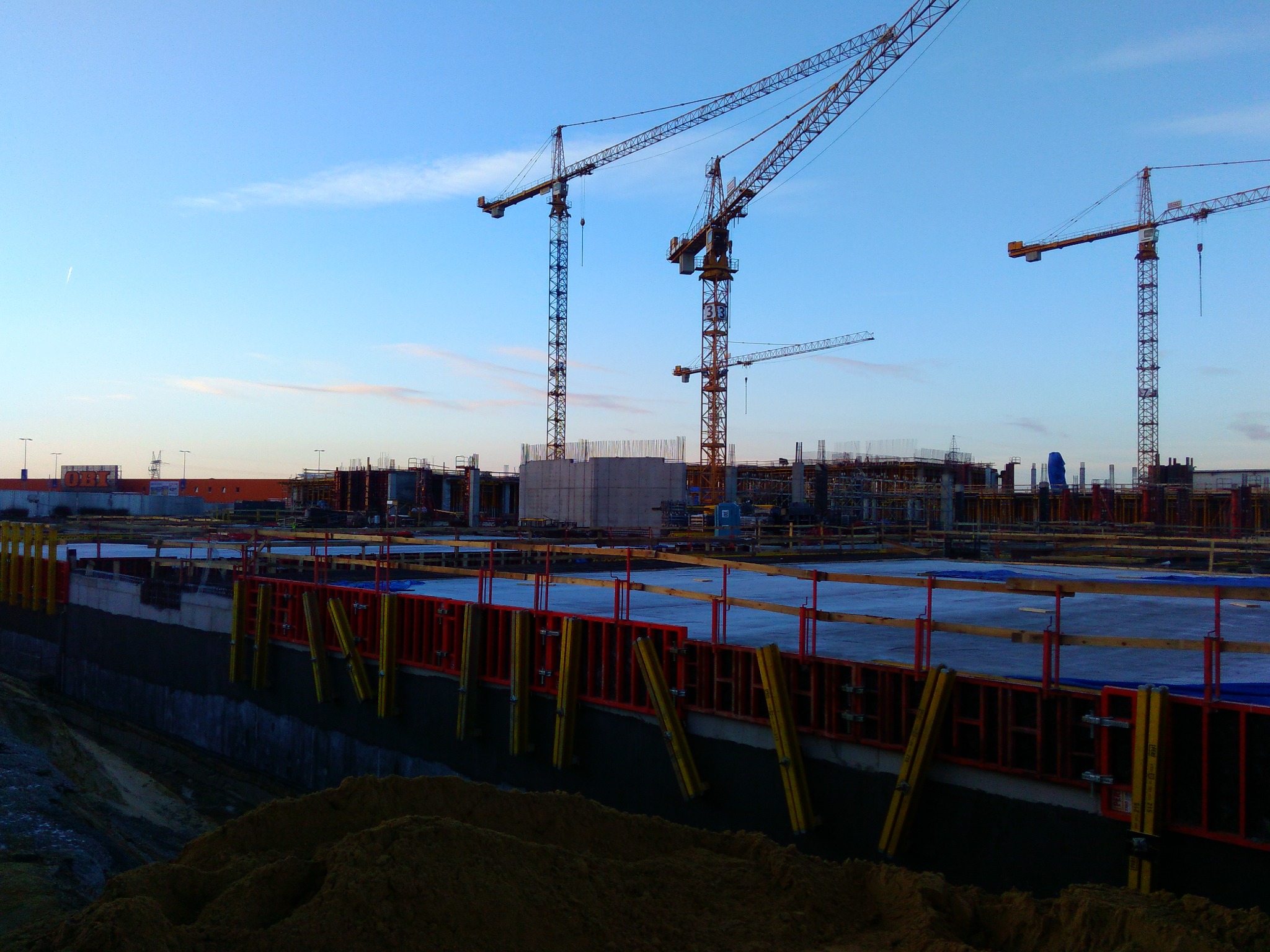 Rozległy plac budowy z widocznymi fundamentami, szalunkami w kolorze czerwonym i żółtym, kilkoma żurawiami budowlanymi na tle błękitnego nieba o zmierzchu.