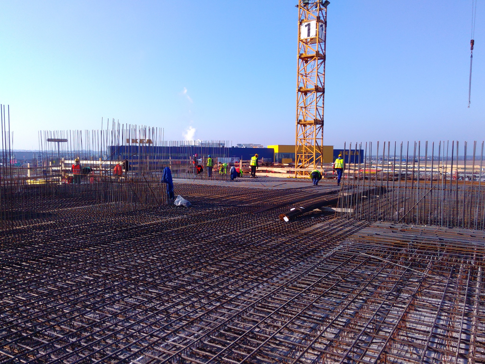 Siatka zbrojeniowa na budowie, pracownicy w kamizelkach odblaskowych, żuraw wieżowy z numerem 1 w tle, widoczny budynek IKEA.