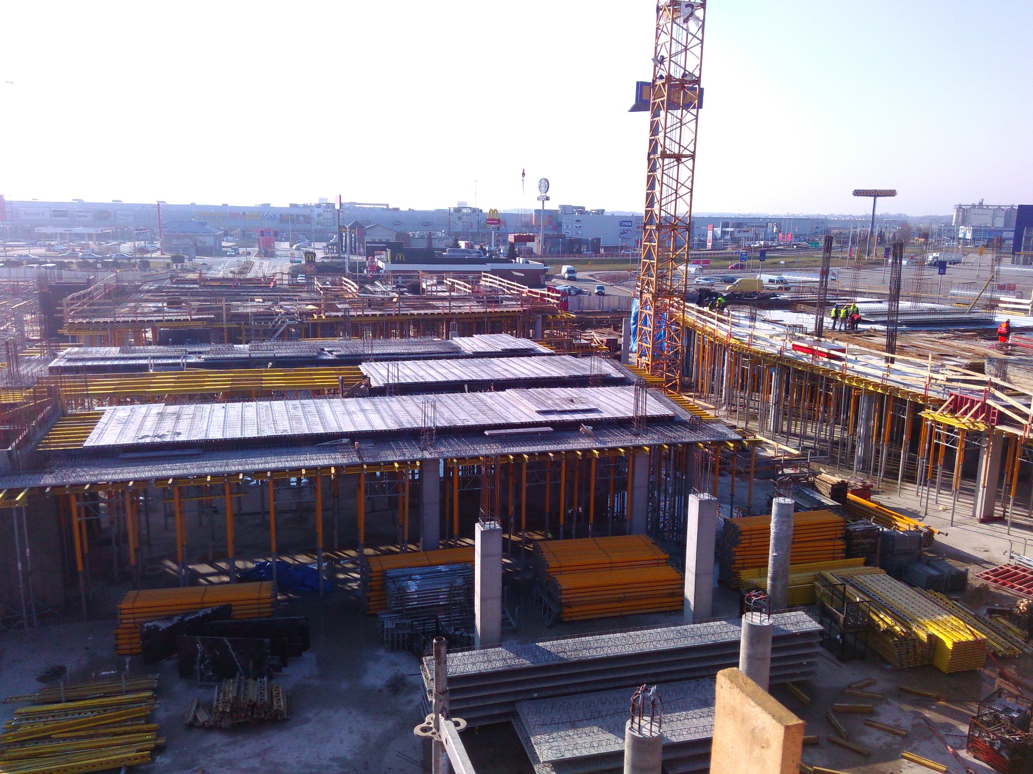 Wysoka perspektywa na plac budowy z widocznymi szalunkami, zbrojeniami i żurawiem wieżowym, w tle zabudowa miejska i rozległe parkingi.