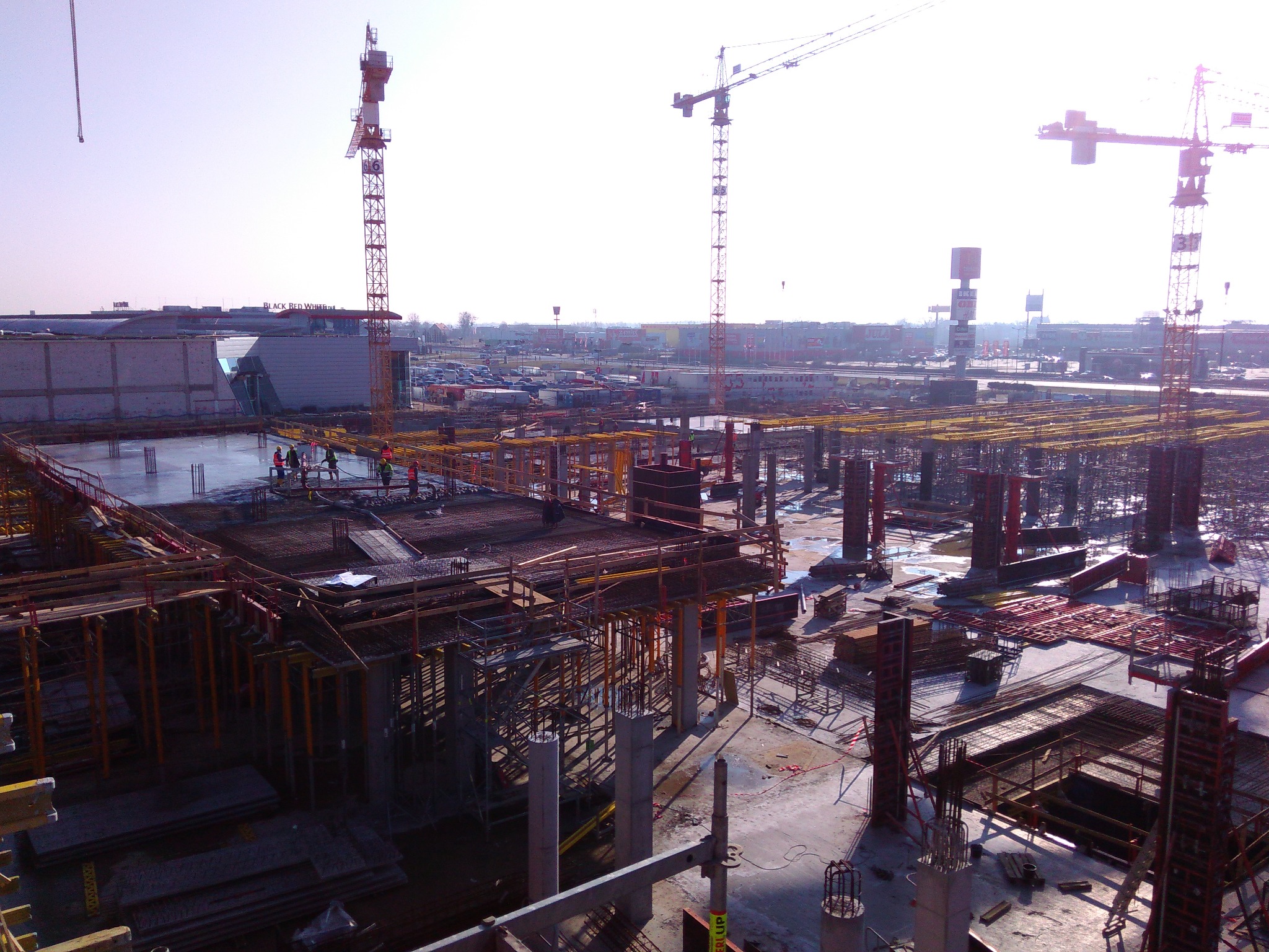 Budowa budynku żelbetowego: fundamenty, szalunki, stalowe zbrojenia, pracownicy w pomarańczowych kamizelkach, liczne żurawie budowlane w tle.