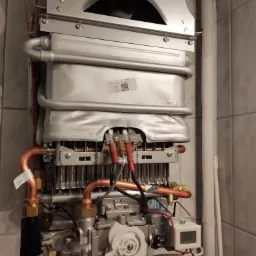 Wewnętrzna budowa pieca gazowego z widocznymi miedzianymi rurami, przewodami elektrycznymi, elementami sterującymi i naklejką z datą produkcji na tle płytek ceramicznych.