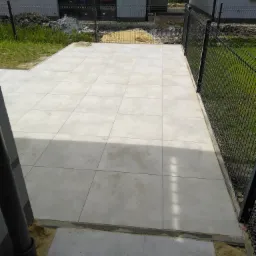 Jasnoszary taras z płytek betonowych, obrzeża wykończone, widoczna czarna metalowa siatka ogrodzeniowa i fragment rynny, teren wokół w trakcie prac ziemnych.