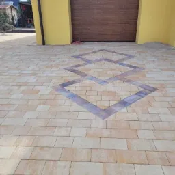 Ułożona kostka brukowa w odcieniach beżu z dekoracyjnym wzorem rombów z ciemniejszych kostek, na podjeździe przed żółtym budynkiem z brązową bramą garażową.