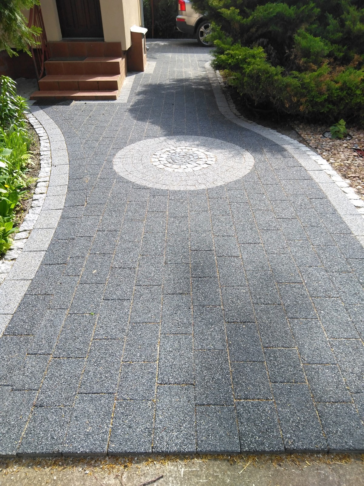 Szara ścieżka z kostki brukowej z dekoracyjnym okręgiem z jasnych kamieni, obrzeża z jasnej kostki, schody wejściowe i roślinność w tle.