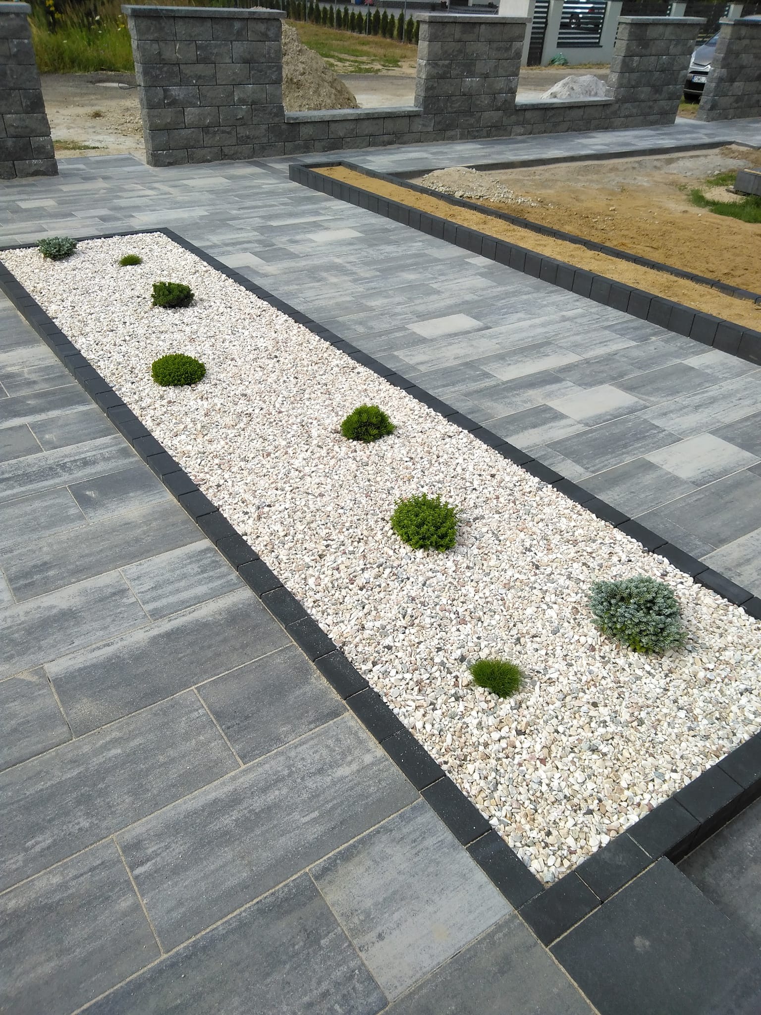 Ułożona szara kostka brukowa z czarną obwódką, wydzielająca pas z białym grysem i nasadzeniami niskich, kulistych krzewów ozdobnych, w tle fragment murku z kostki.