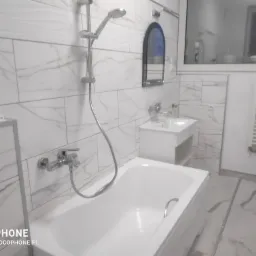 Nowoczesna, biała łazienka z wanną, umywalką i marmurowymi płytkami na ścianach i podłodze, lustro w czarnej ramie, grzejnik drabinkowy.