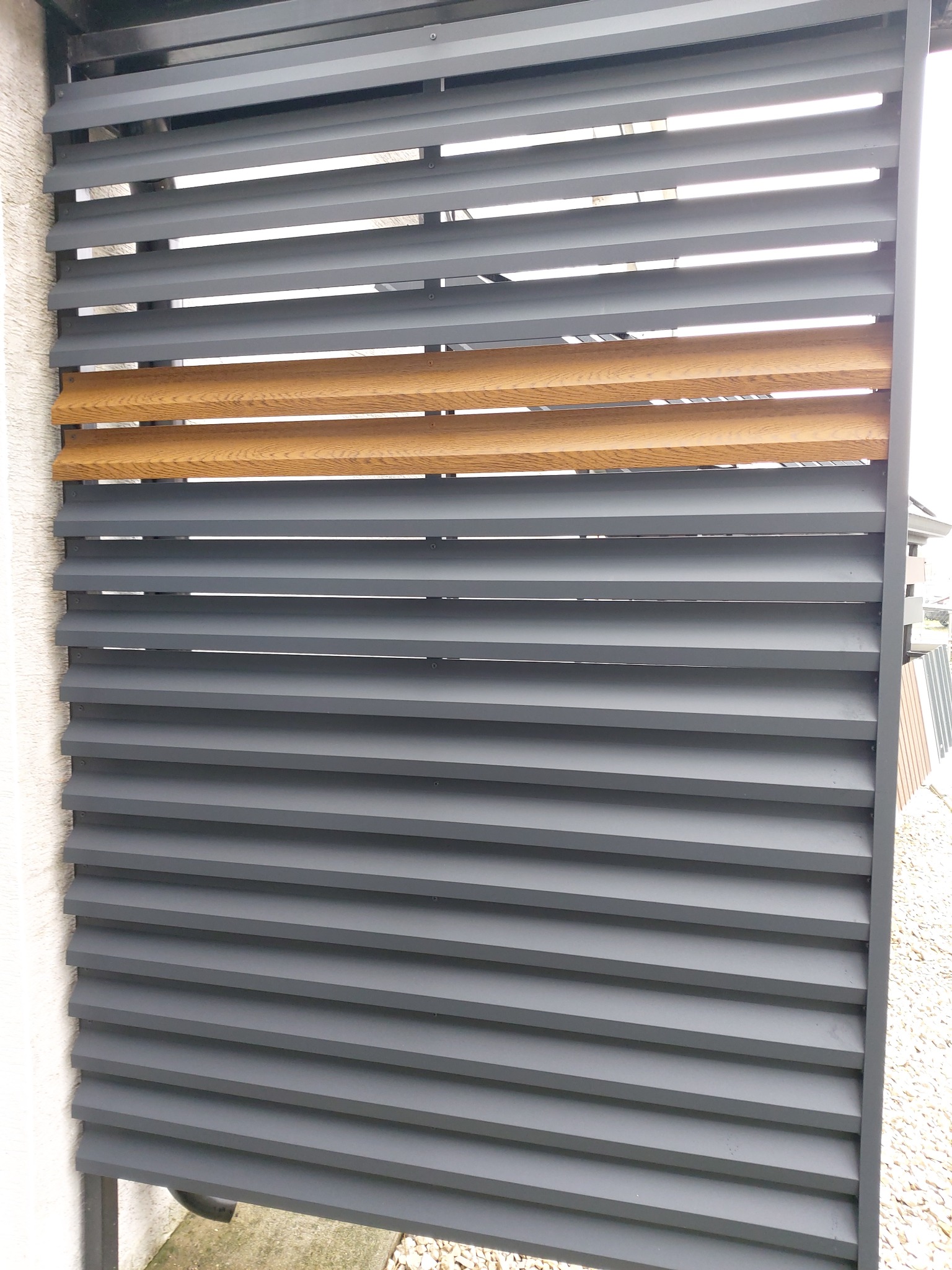 Nowoczesne ogrodzenie z szarych paneli z pojedynczym rzędem drewnianych desek w kolorze jasnego dębu, widoczne z bliska, z fragmentem elewacji budynku po lewej stronie.