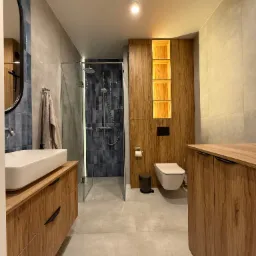 Nowoczesna łazienka z drewnianymi szafkami, umywalką nablatową, kabiną prysznicową ze szklaną ścianą i toaletą podwieszaną z podświetlanymi wnękami w zabudowie.