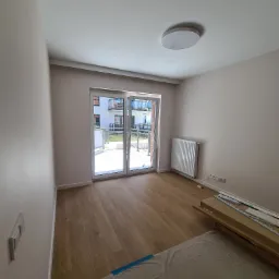 Pusty pok&oacute;j po remoncie z drewnianą podłogą, kremowymi ścianami, grzejnikiem, oknem balkonowym z widokiem na osiedle i rozłożoną folią ochronną na podłodze przykrywającą materiały budowlane.