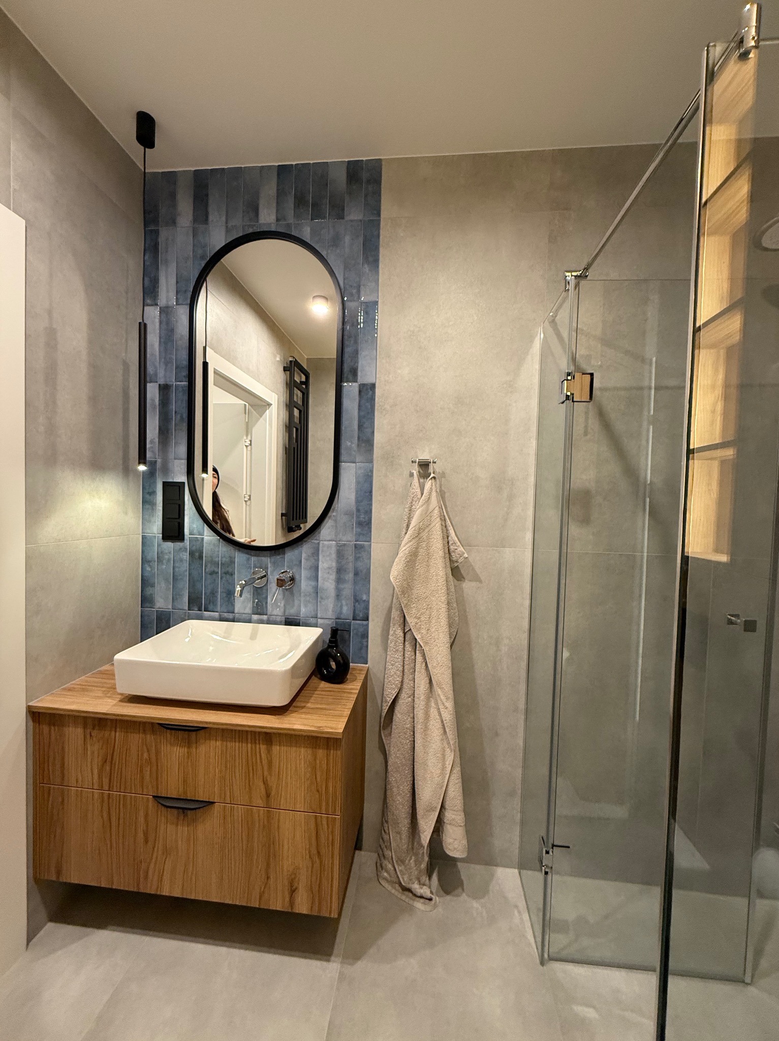 Nowoczesna łazienka z drewnianą szafką pod umywalkę, owalnym lustrem z czarną ramą, niebieskimi płytkami na ścianie, szklaną kabiną prysznicową i beżowym ręcznikiem.