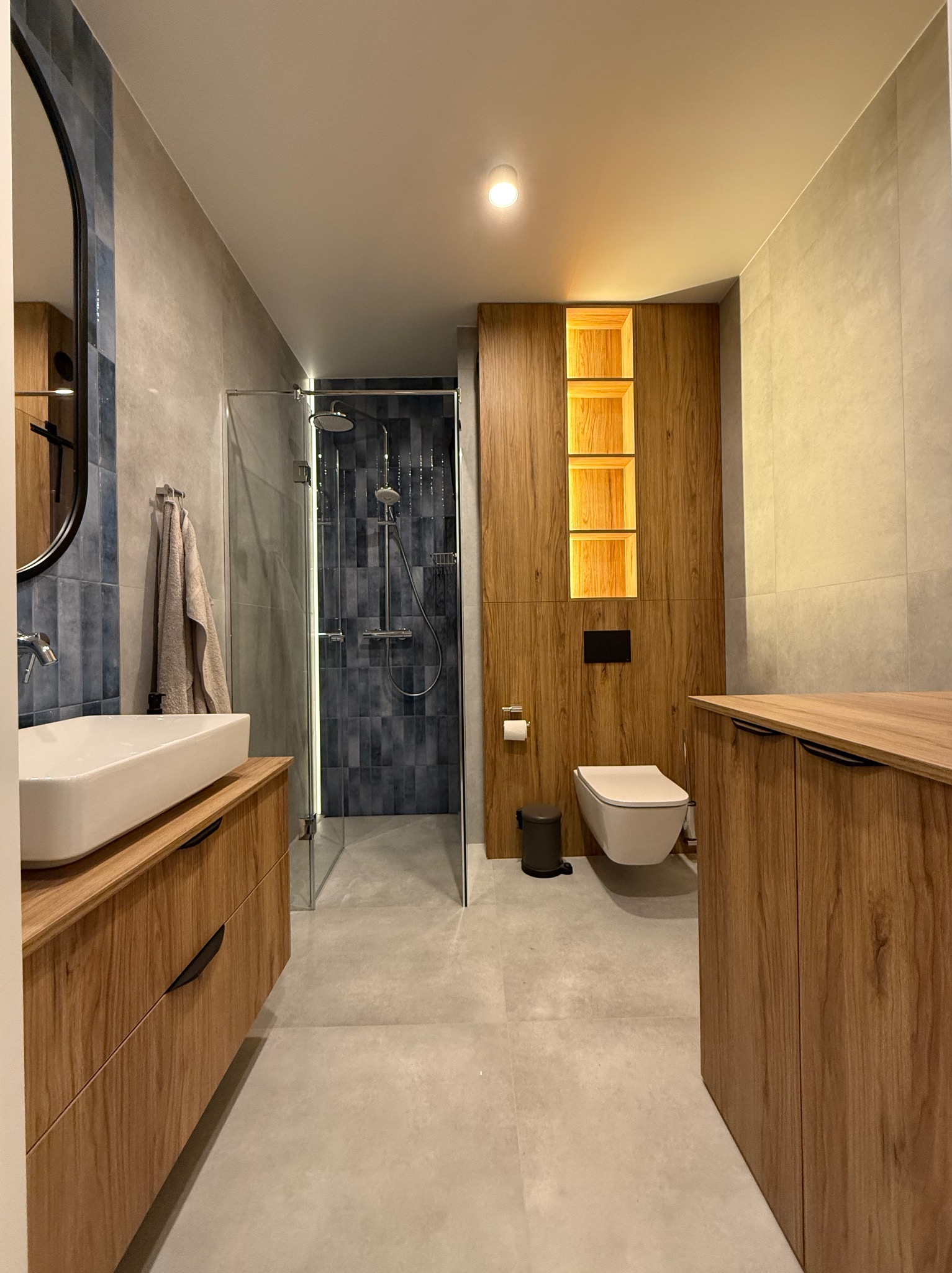 Nowoczesna łazienka z drewnianymi szafkami, umywalką nablatową, kabiną prysznicową ze szklaną ścianą i toaletą podwieszaną z podświetlanymi wnękami w zabudowie.