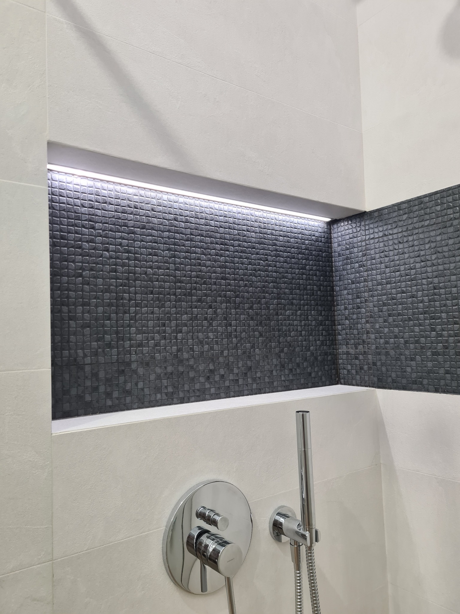 Nowoczesna łazienka z wnęką wyłożoną ciemną mozaiką i oświetleniem LED, chromowana bateria natryskowa na jasnych płytkach.