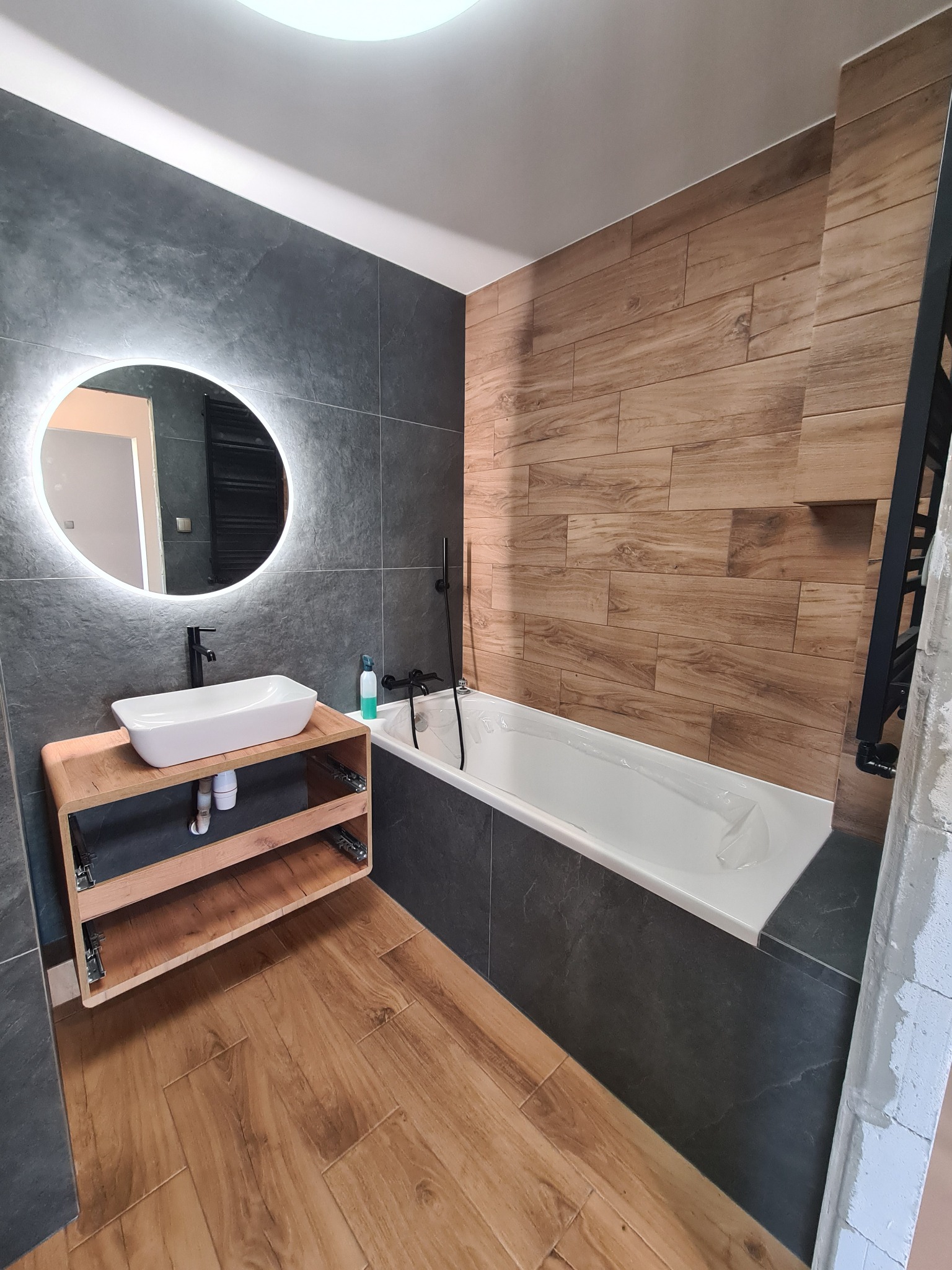 Nowoczesna łazienka z drewnopodobnymi płytkami na podłodze i ścianie, szare płytki imitujące kamień, okrągłe lustro z podświetleniem LED nad umywalką na drewnianej szafce, biała wanna z czarną...