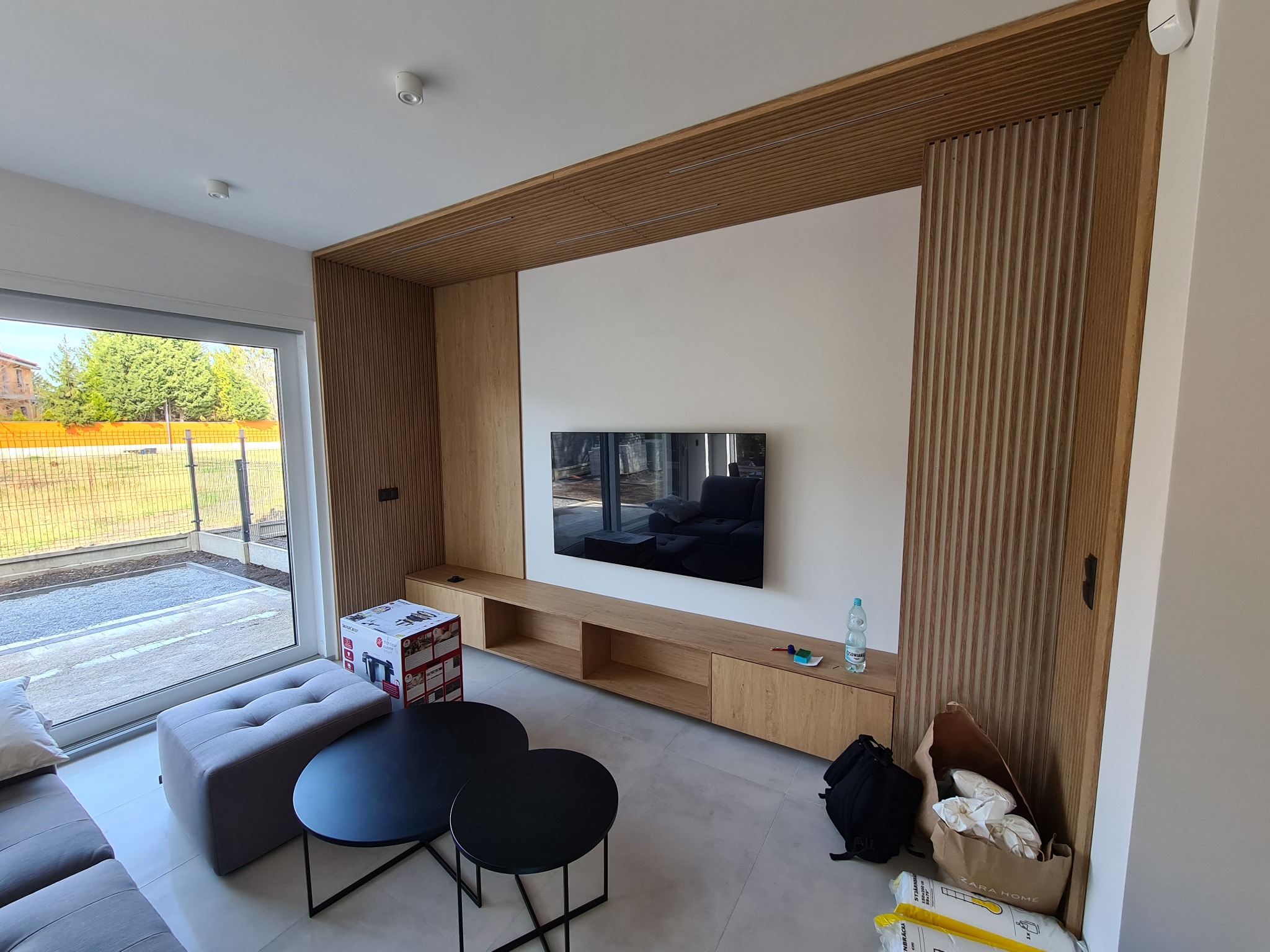 Salon z drewnianą zabudową ściany telewizyjnej, widoczne meble, telewizor i elementy dekoracyjne, okno z widokiem na zewnątrz. Na podłodze kartony i torba.