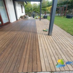 MP clean - Drewniany taras po czyszczeniu: połowa desek w ciemnym odcieniu, druga połowa w jasnym. Widoczna szczotka do czyszczenia. Tło: trawnik, dom.