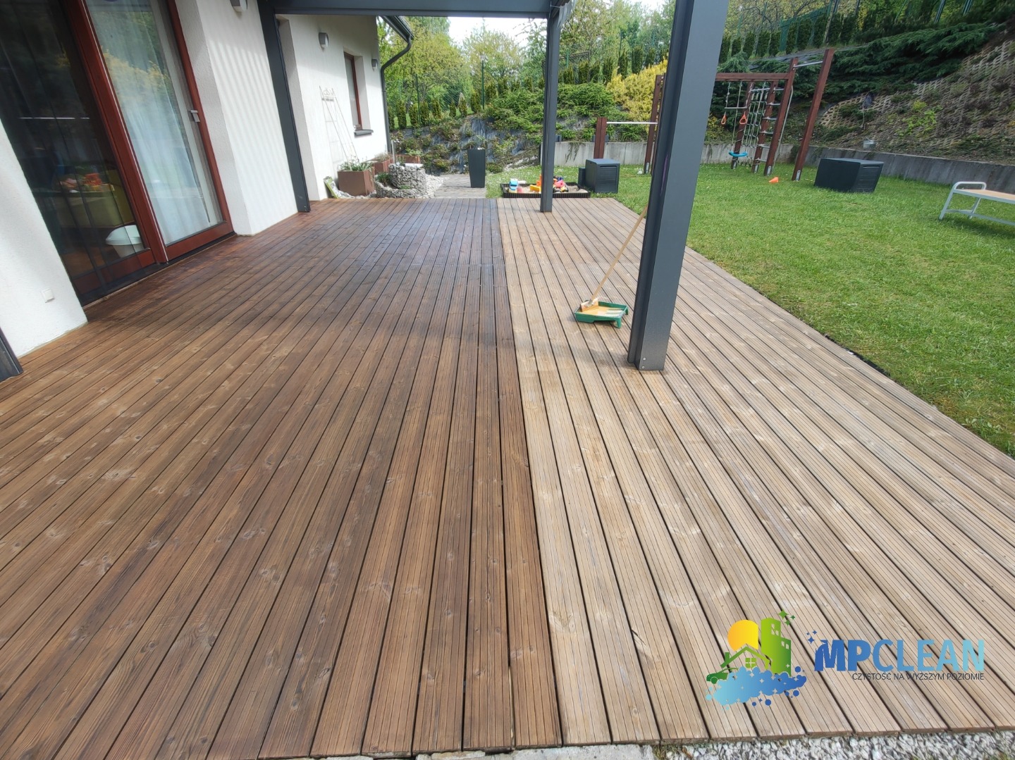 Drewniany taras po czyszczeniu: połowa desek w ciemnym odcieniu, druga połowa w jasnym. Widoczna szczotka do czyszczenia. Tło: trawnik, dom.