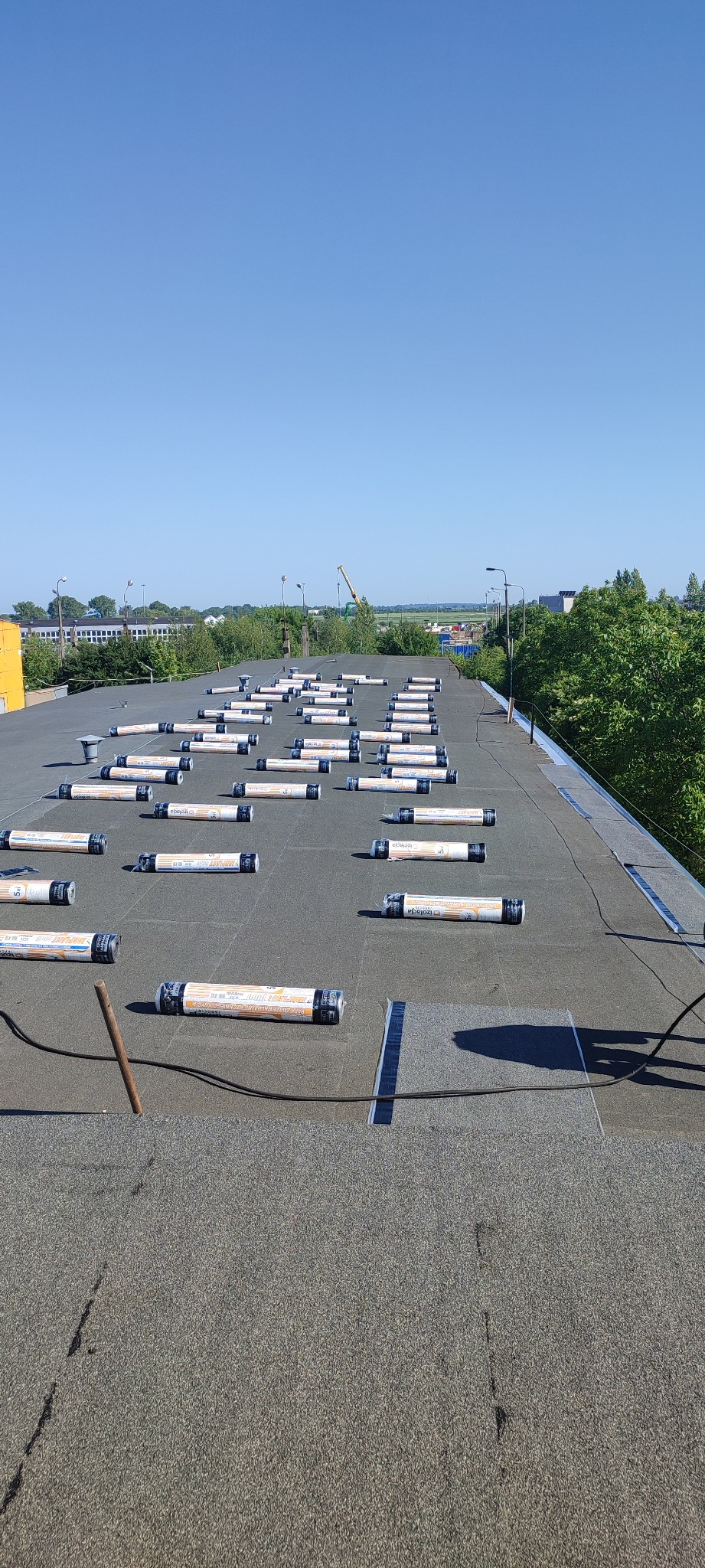 Widok z góry na płaski dach pokryty papą, na którym rozmieszczono rolki papy przygotowane do montażu, z widocznymi fragmentami taśmy uszczelniającej i elementami krajobrazu w tle.