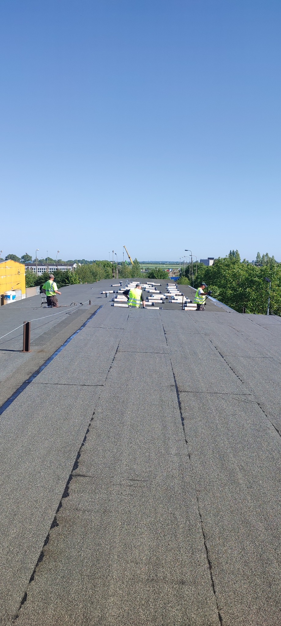 Pracownicy w jaskrawych kamizelkach montują konstrukcję wsporczą pod panele słoneczne na płaskim dachu pokrytym papą. W tle widoczny krajobraz miejski i błękitne niebo.