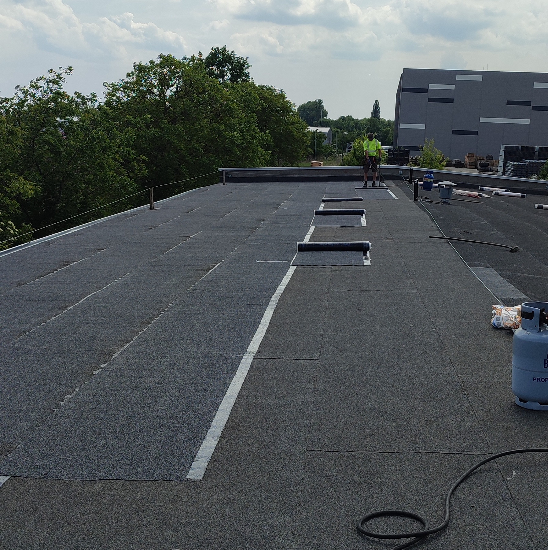 Prace dekarskie na dachu płaskim, widoczny dekarz w odblaskowej kamizelce, rolki papy, butla z gazem i zgrzewarka.