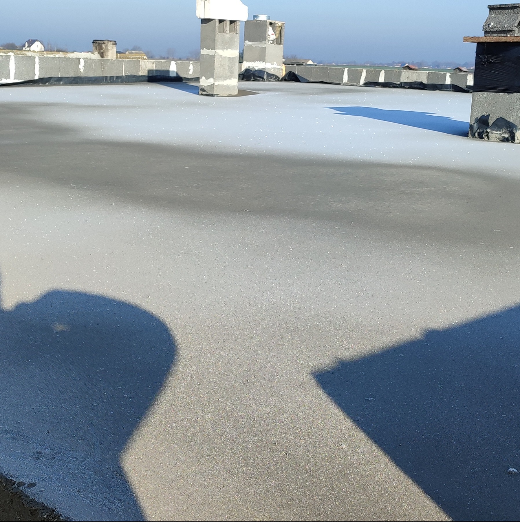 Wykończony płaski dach pokryty szarą hydroizolacją, z widocznymi kominami wentylacyjnymi z szarego betonu komórkowego i obróbkami blacharskimi na krawędziach, w tle zabudowa wiejska.