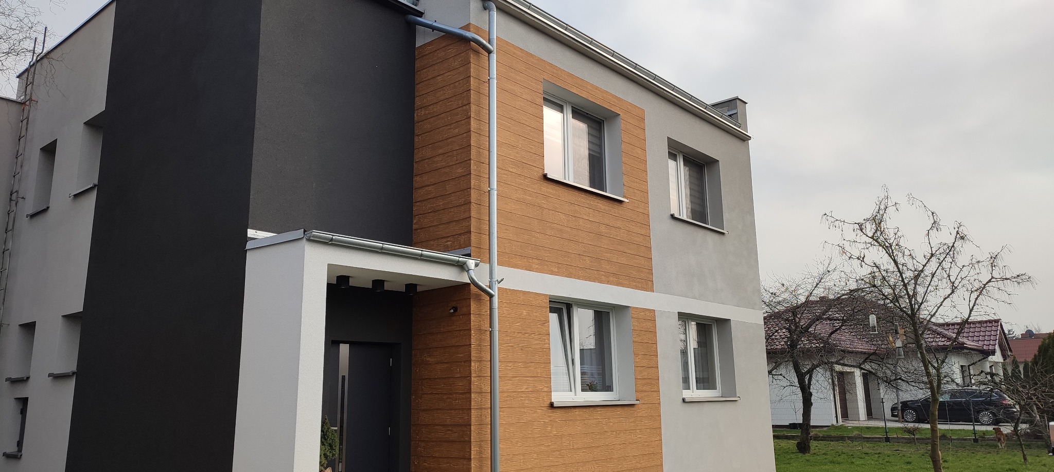 Nowoczesna elewacja dwukondygnacyjnego domu z drewnopodobną okładziną, szarą ścianą i białym wejściem z widocznymi rynnami.