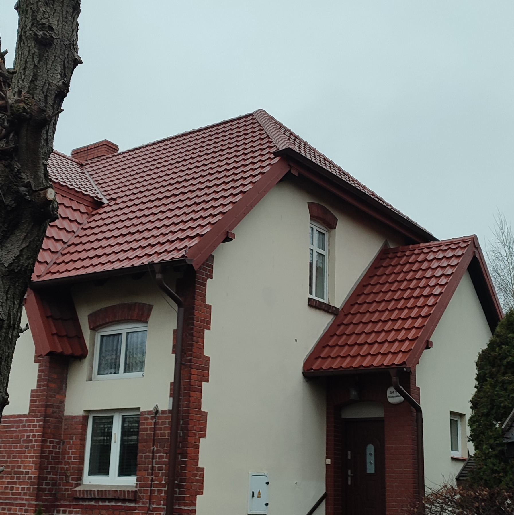 Dwupiętrowy dom z nową, czerwoną dachówką karpiówką i ceglaną elewacją, widoczny z poziomu ulicy w pochmurny dzień.