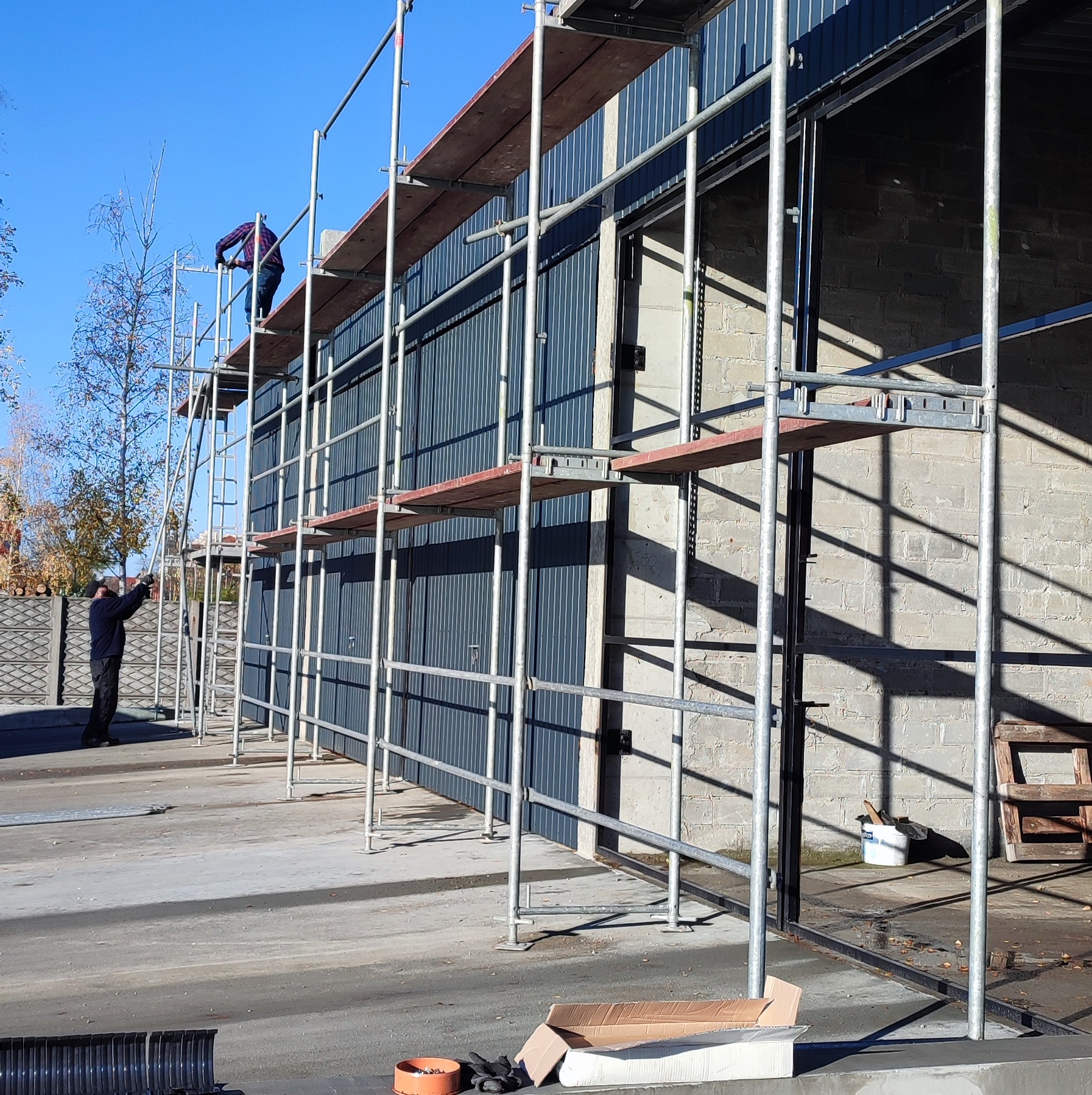 Pracownicy na rusztowaniu montują blaszane poszycie na budynku z cegły, widoczne narzędzia i materiały budowlane na ziemi.